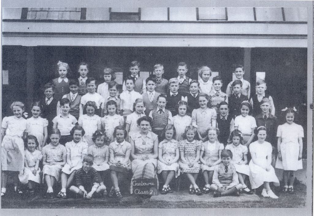 A class of about forty-five children in rows with a woman teacher seated in the middle, the girls in summer dresses; a slate at the front reads “Cranbourne Class 2”.