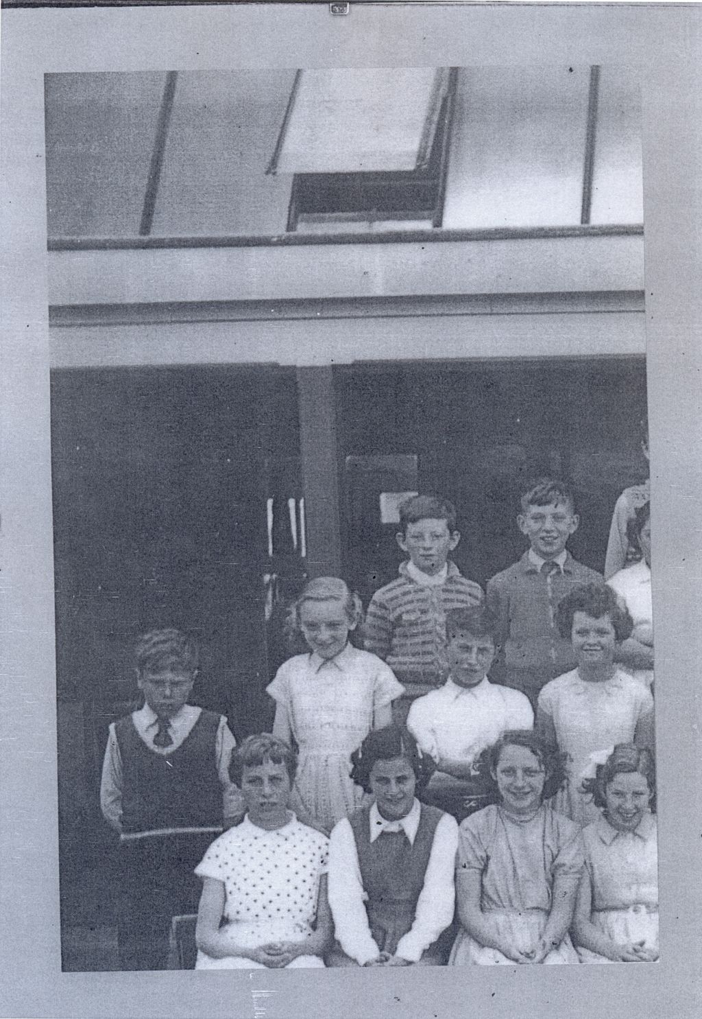 Part of a class photograph mounted on a grey album page: about a dozen children in three rows in front of a verandah and roof light, the rest of the class cut off at the left.