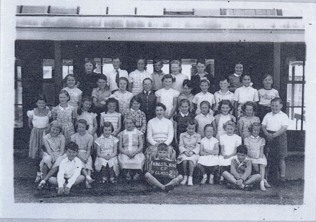 A class of about forty children in rows with a woman teacher in the centre, girls in patterned summer dresses; a boy at the front holds a slate reading “Kingsthorne C.P. Class 2”.