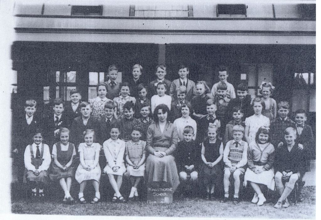 A class of about forty children in rows in front of a verandah, with a woman teacher seated in the centre; a slate at her feet reads “Kingsthorne School”.