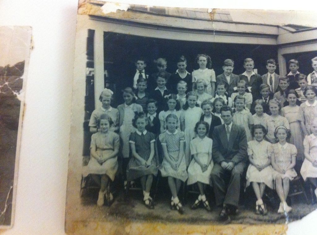 A photograph of a school photograph, partly torn at the corners: a class of about forty boys and girls in rows in front of a verandah, with a male teacher in a suit seated in the middle of the front row. The edge of another print shows at the left.