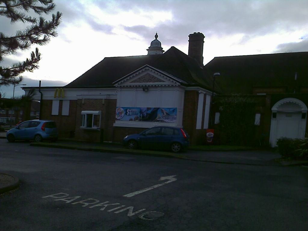 The former College Arms pub, a long brick building with a tiled roof, a small cupola and a white pedimented entrance, now carrying McDonald’s golden arches and a promotional banner; cars are parked in front and “PARKING” is painted on the road.