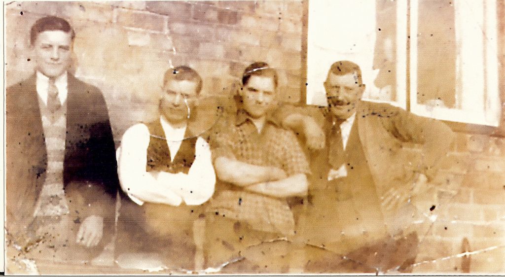 A faded, spotted sepia photograph of four men sitting against a brick wall beside a window: a young man in a suit and patterned pullover, two men in shirtsleeves and waistcoats, and an older moustached man in a jacket at the right.