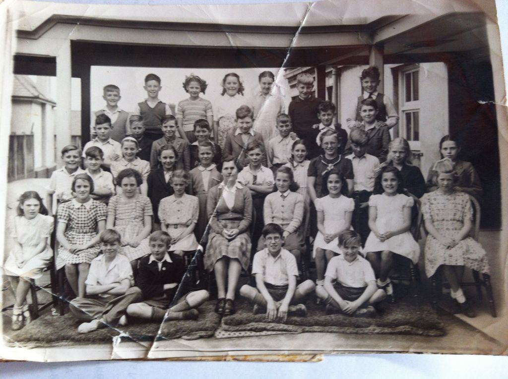 A creased and cracked class photograph: about thirty-five children in four rows under a verandah, the boys at the front sitting cross-legged on mats and a woman teacher seated in the centre.