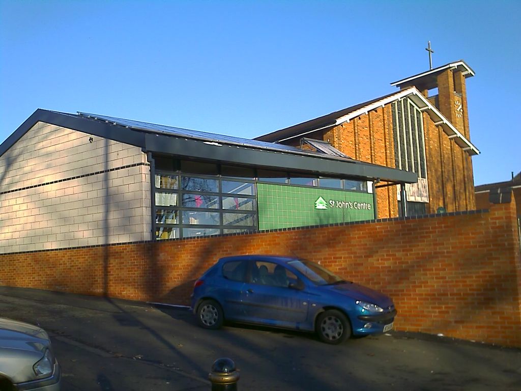 A modern single-storey community building with a green-tiled front lettered “St John’s Centre” stands beside the brick gable and clock tower of Christ the King church, behind a new brick wall; a blue car is parked in the road.