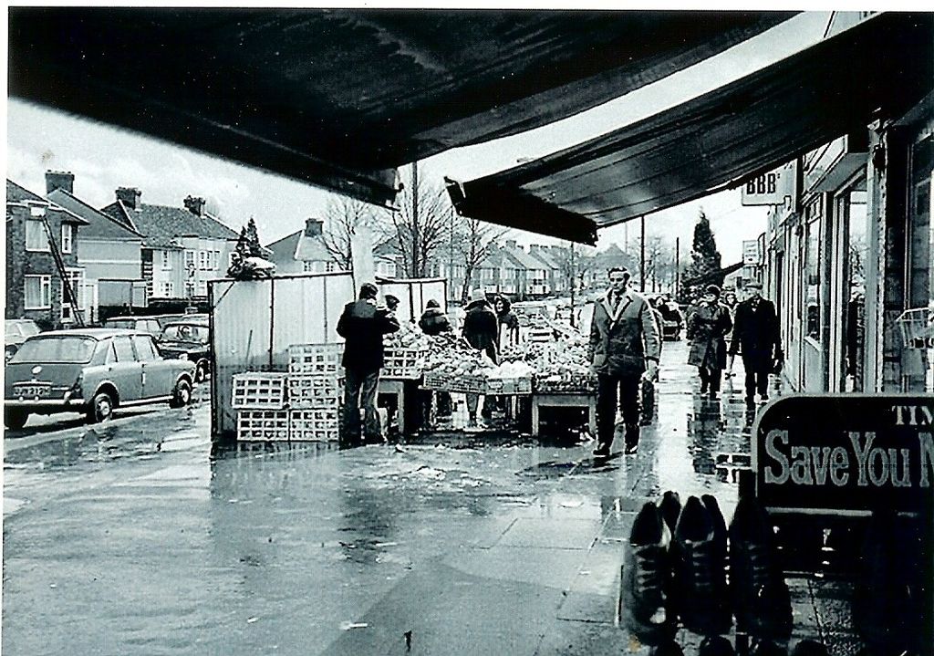 Looking along a rain-soaked pavement from under shop awnings: a greengrocer’s stall piled with produce and crates, shoppers in coats, a parked 1960s saloon car, semi-detached houses across the road and a shoe-shop sign reading “Save You M…” in the foreground.