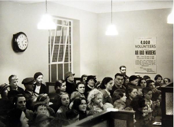 A crowd of adults and children packs the banking hall behind a wooden counter, watching the ceremony; a wall clock hangs on the left and a poster on the right appeals for “4,000 Volunteers required as Air Raid Wardens”.