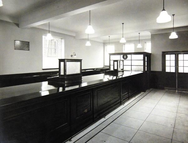 The empty banking hall of the new branch: a long polished wooden counter with a glazed screen, a tiled floor, pendant lamps, a plaque on the wall and a partly glazed office at the far end.