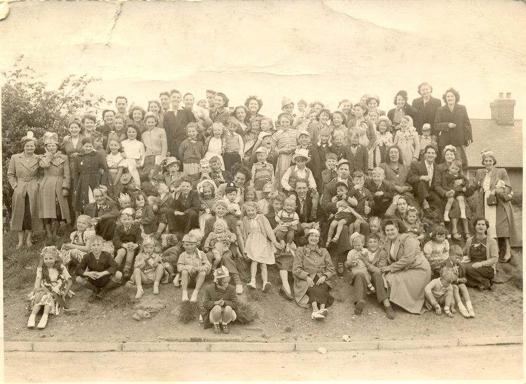 A large crowd of neighbours – women in coats and hats, men, and children in paper crowns and fancy dress – posed on a grassy bank above a kerb, with a house chimney at the right.
