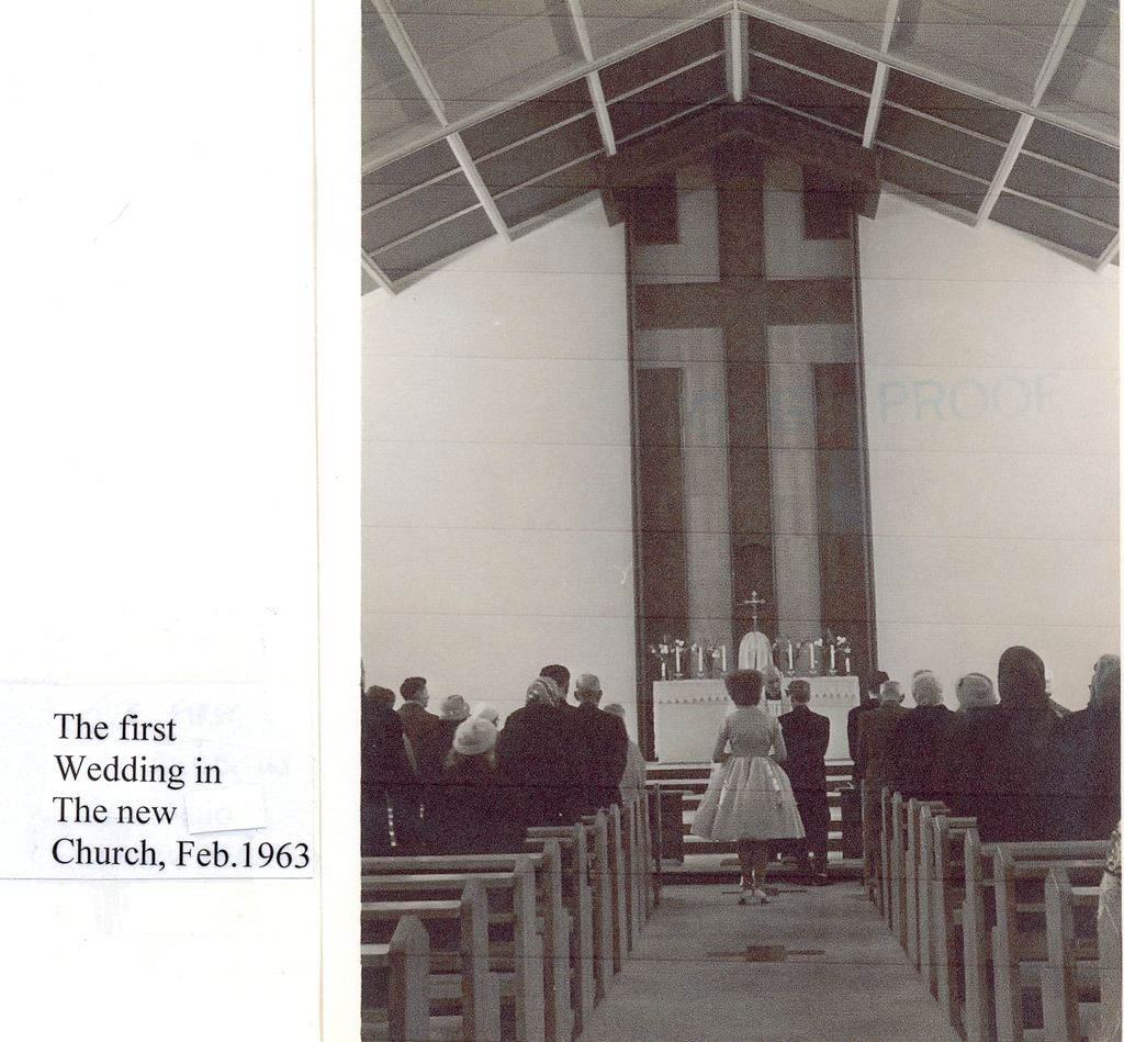 The interior of the new church, looking down the aisle between rows of pews to a bride and groom at the altar under a tall timber cross and a steep boarded roof; a label reads “The first Wedding in The new Church, Feb. 1963”.