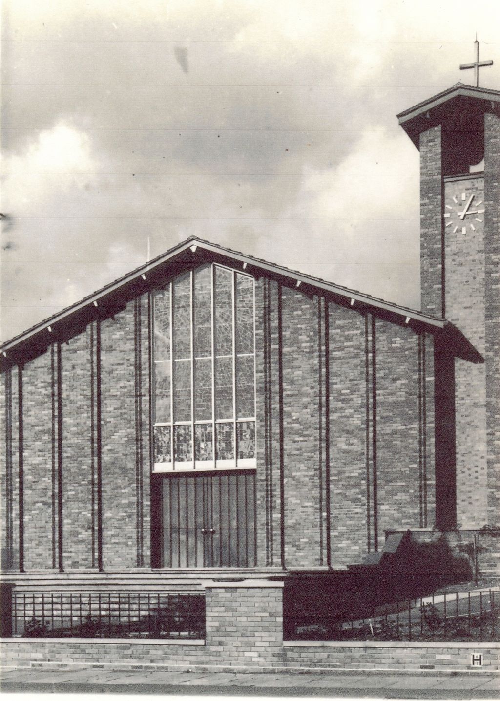 A black-and-white view of the new church’s brick west front: a tall gable with a large stained-glass window above double doors, a clock tower with a cross on the right, and a low wall with railings in front.