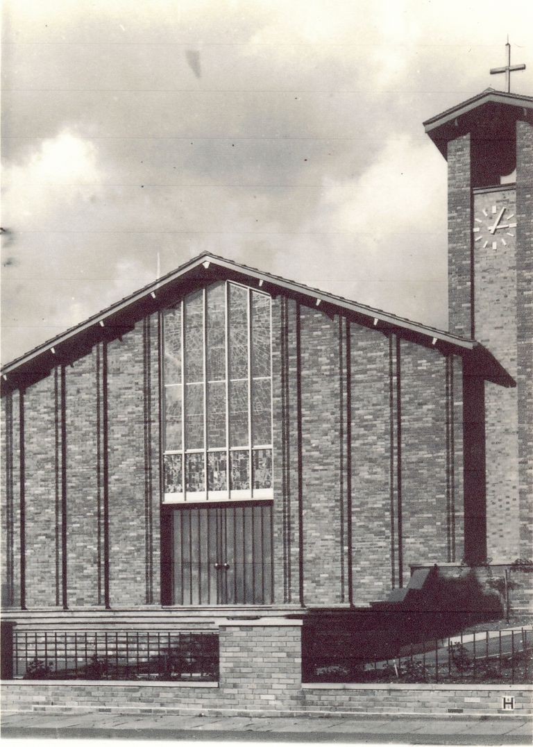 A black-and-white view of the new church’s brick west front: a tall gable with a large stained-glass window above double doors, a clock tower with a cross on the right, and a low wall with railings in front.