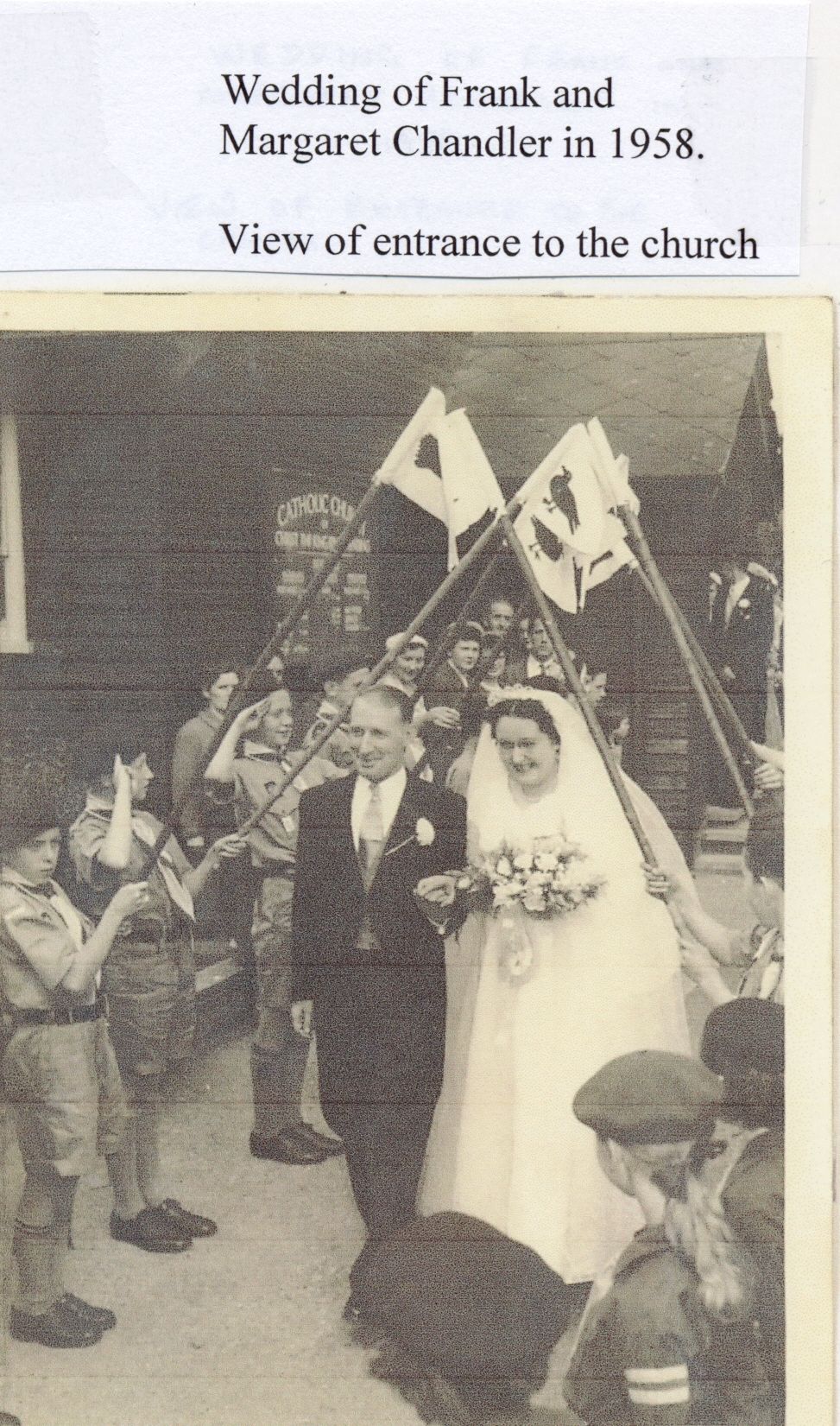 A bride and groom leave a wooden church under an arch of flags on staves held by scouts and cubs; a noticeboard behind reads “Catholic Church of Christ the King”. A label above says “Wedding of Frank and Margaret Chandler in 1958. View of entrance to the church”.