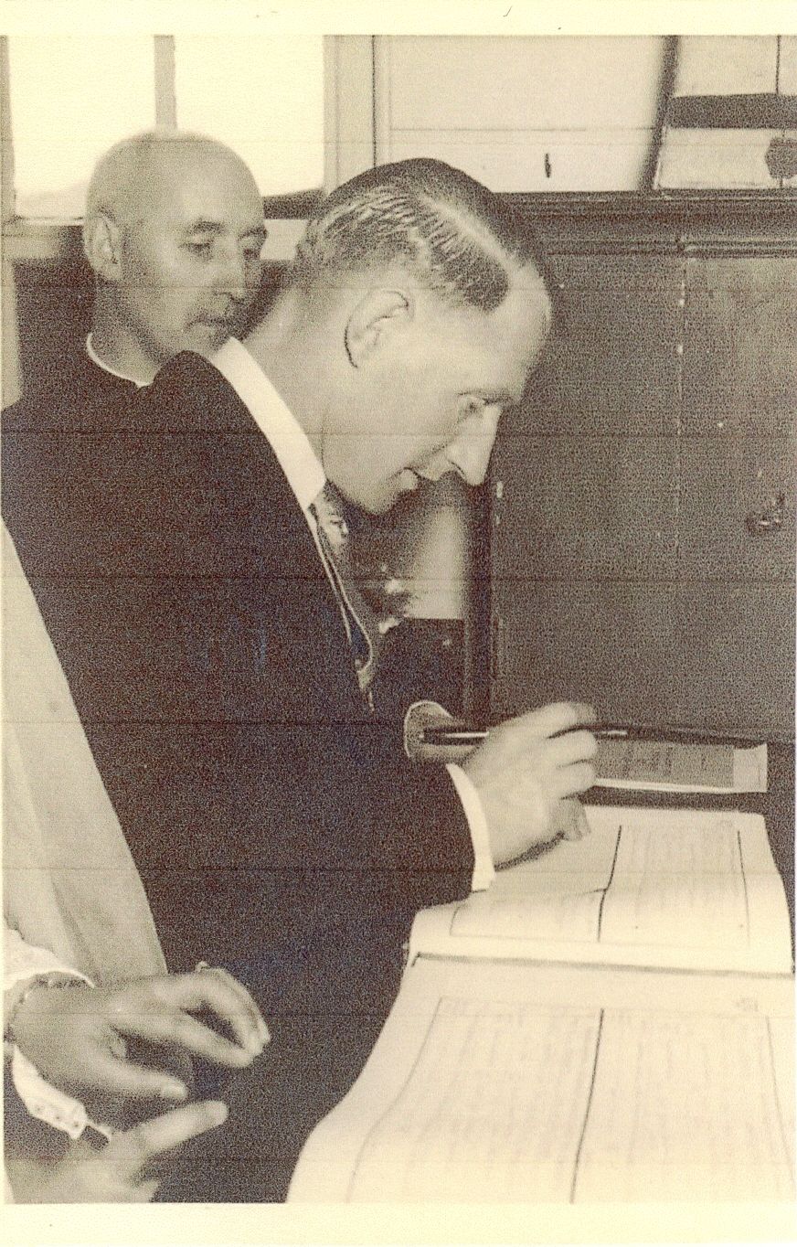 A sepia close-up of a man in a suit signing a large register at a table while a priest looks on from behind; a bride’s hand is visible at the lower left.