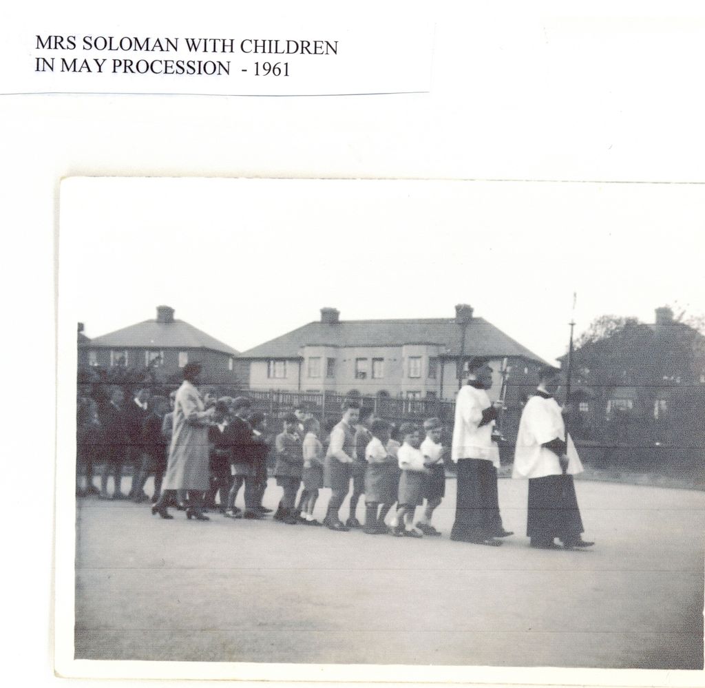 A line of small children led by two altar servers in cassocks and surplices, one carrying a cross, walks across a playground with a woman teacher, with 1930s semi-detached houses behind; the label reads “Mrs Soloman with children in May procession – 1961”.