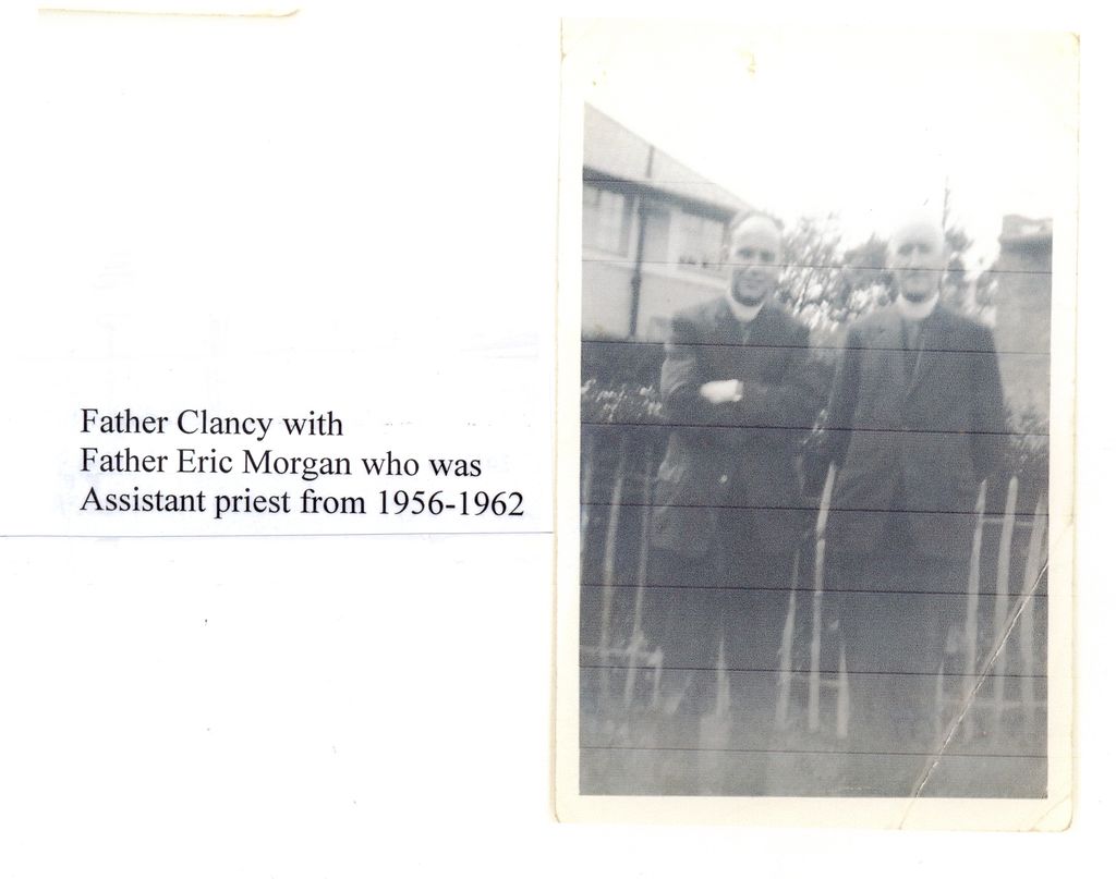 A faded snapshot of two priests in dark suits and collars standing in a garden by a fence, with houses behind; the label reads “Father Clancy with Father Eric Morgan who was Assistant priest from 1956-1962”.