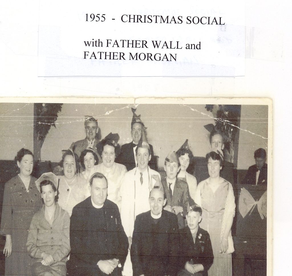 A creased group photograph of about sixteen parishioners in paper hats at a Christmas party, with two priests seated in the front row; the typed label reads “1955 – Christmas social with Father Wall and Father Morgan”.