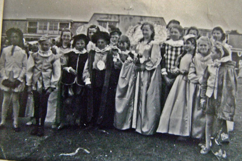 Children in Tudor-style pageant costume – ruffs, long satin gowns, a crowned “queen” with a large fan-like collar – stand on grass in front of the school’s flat-roofed buildings.