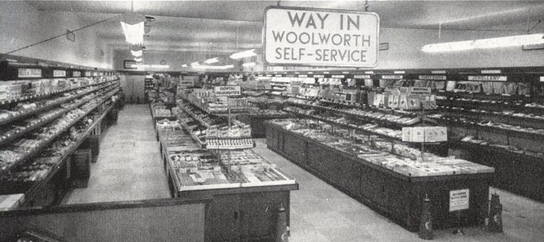 The inside of a Woolworth store: long island counters and wall shelves stacked with goods, strip lights overhead and a hanging sign reading “Way in – Woolworth self-service”.