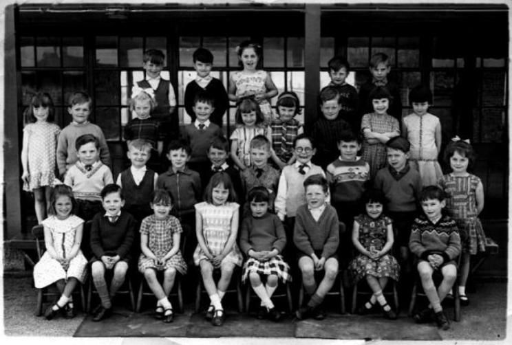 A class of about thirty-five children in four rows, the front row seated on chairs with a mat at their feet, in front of a glazed classroom wall.