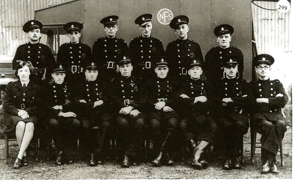 Fourteen firemen in double-breasted tunics, belts and peaked caps and one woman officer in uniform, posed in two rows in front of a hut marked “NFS”; a lorry stands at the left. A circled “299” is printed in the top corner.