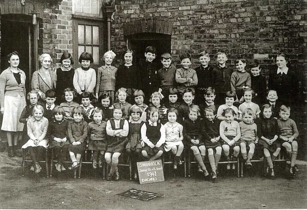 A school group of about forty children in three rows, with three women teachers, in front of a brick school building. A slate on the ground reads “Swanwick … 1941 Evacuees”.