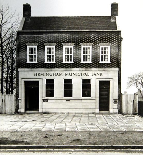 A newly built two-storey brick bank with sash windows, chimneys at each end and a pale stone ground floor lettered “Birmingham Municipal Bank”, with two doors, a paved forecourt and a wooden fence on either side.