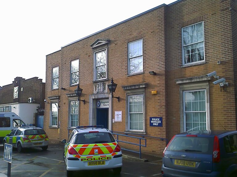 The two-storey brick police station with a pedimented doorway flanked by blue police lamps, and marked police cars parked in the yard in front.