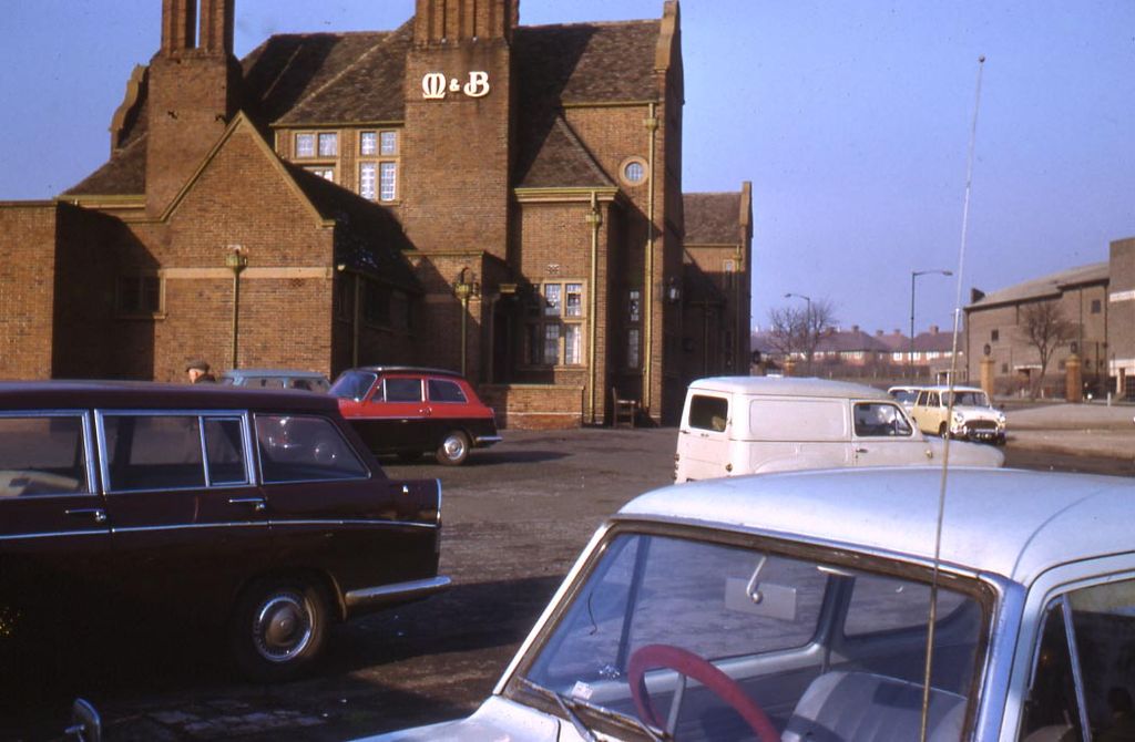 Colour photograph of a large brick pub with tall chimneys and an “M&B” sign on its gable, with 1960s cars and a van parked on the forecourt and houses in the distance.