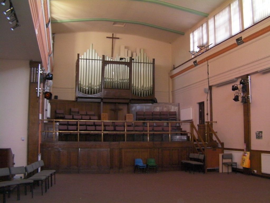 The inside of a tall church hall with a large pipe organ and a wooden cross above tiered choir seating behind a brass rail, stage lights on the walls and rows of chairs on the carpet.