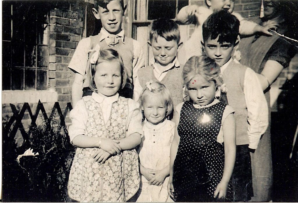 Black-and-white snapshot of three young girls in summer frocks standing in front of three older boys in knitted tank tops, outside a brick house with a trellis fence; an adult and a baby are partly visible behind.