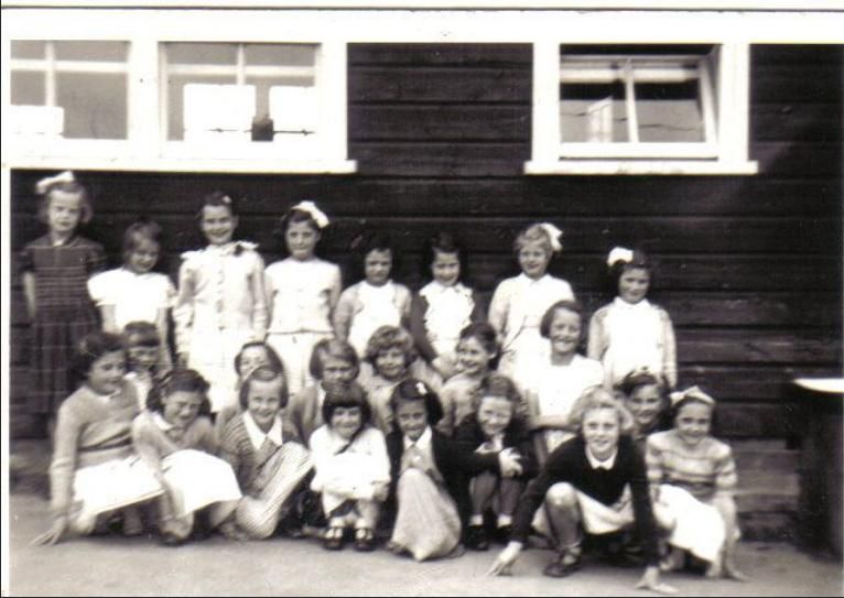 Black-and-white group photograph of about twenty young girls, many with ribbons in their hair, in two rows in front of a dark timber classroom hut with white-framed windows.