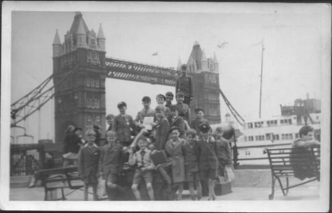 Black-and-white snapshot of a group of schoolboys in caps and short trousers crowded on and around a bench on the riverside, with Tower Bridge behind them and a pleasure boat on the right.