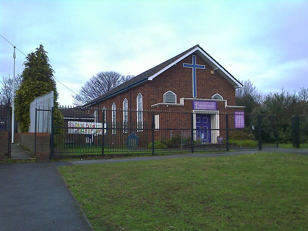 A red-brick gabled church with a large blue cross on its front, arched windows and a purple “The Link Church” sign, behind black railings with a playgroup banner at the side.