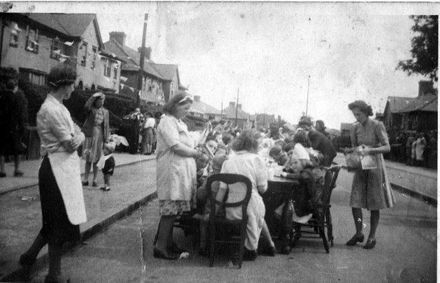 Black-and-white photograph of long tables down the middle of a street of semi-detached houses, crowded with children, while women in aprons and headscarves serve and watch.