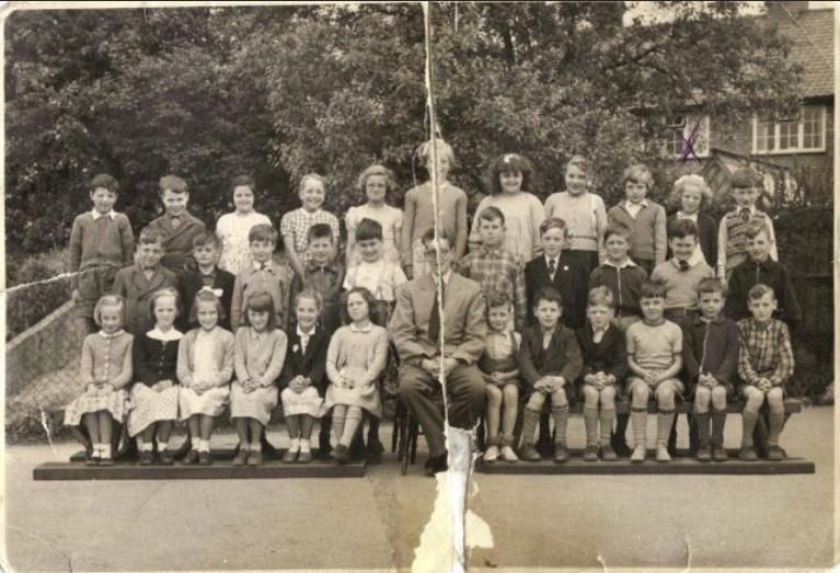 Black-and-white class photograph of about thirty-five children in three rows on benches in a school yard, with a male teacher in a suit seated in the middle; the print is torn and creased.
