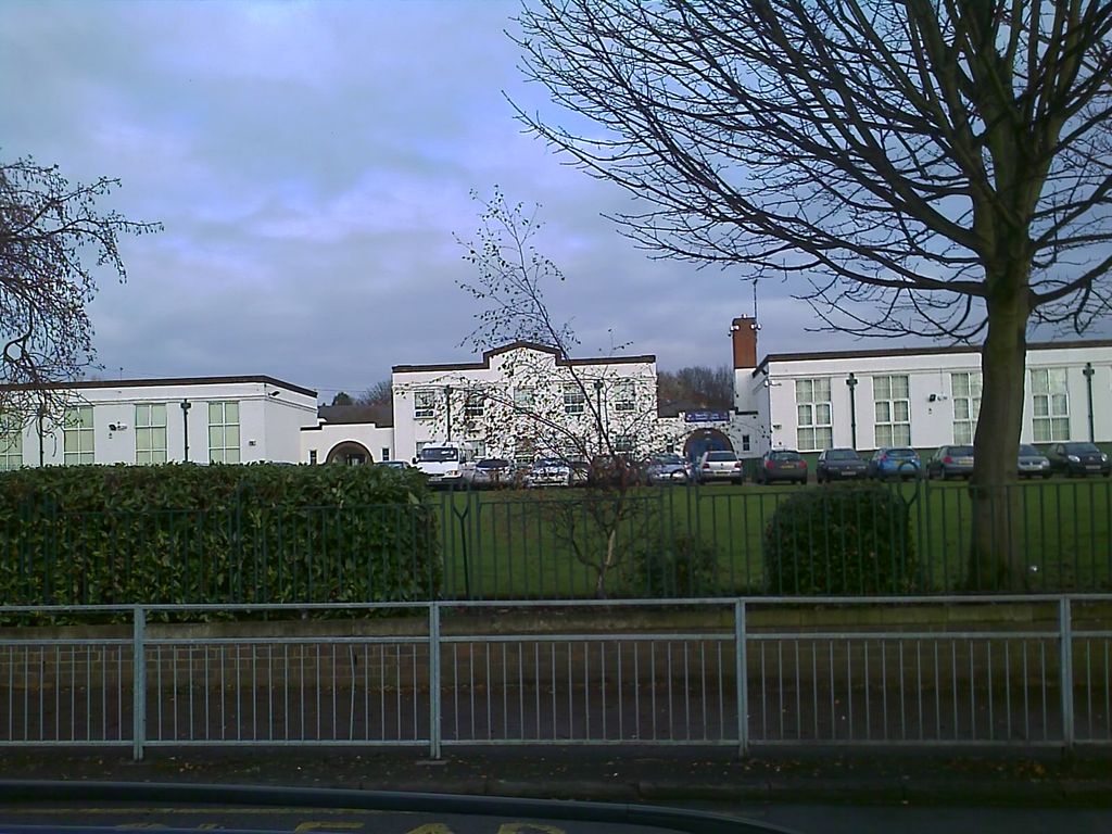 A long, white-rendered school building with a stepped central gable, a tall brick chimney and green-framed windows, seen across a playing field behind railings and a hedge.