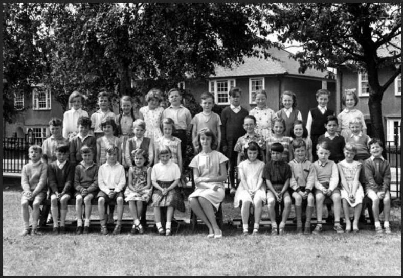 Black-and-white class photograph of about thirty children in three rows on benches on a lawn, a young woman teacher seated in the centre, with semi-detached houses and trees behind the railings.