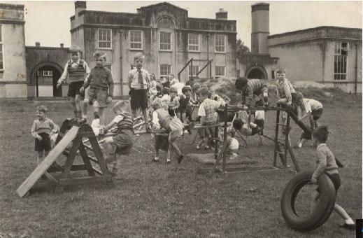 Young boys play on wooden PE apparatus on the school field – sloping ladders, a climbing frame and a tyre – in front of a two-storey school building with a tall chimney.