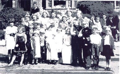 Small black-and-white group photograph of children in fancy dress and party hats standing on a pavement in front of houses and a hedge, with adults behind.