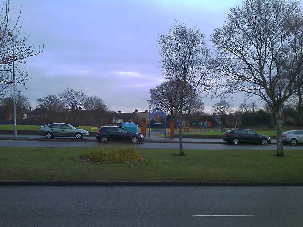 A grassed park with a children’s playground behind a blue arched gateway between brick pillars, seen across a road and a grass verge with young trees; houses line the far side.
