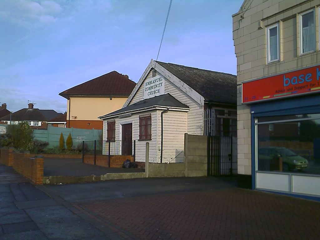 A small white weatherboarded chapel with a gabled front and a sign reading “Emmanuel Community Church”, beside a modern shopfront with a red sign, behind a low brick wall.