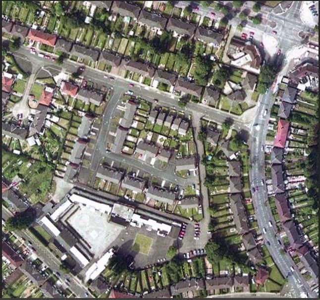 A vertical colour aerial view of streets of semi-detached houses with long back gardens and parked cars, with a flat-roofed school building and playground at the bottom left.