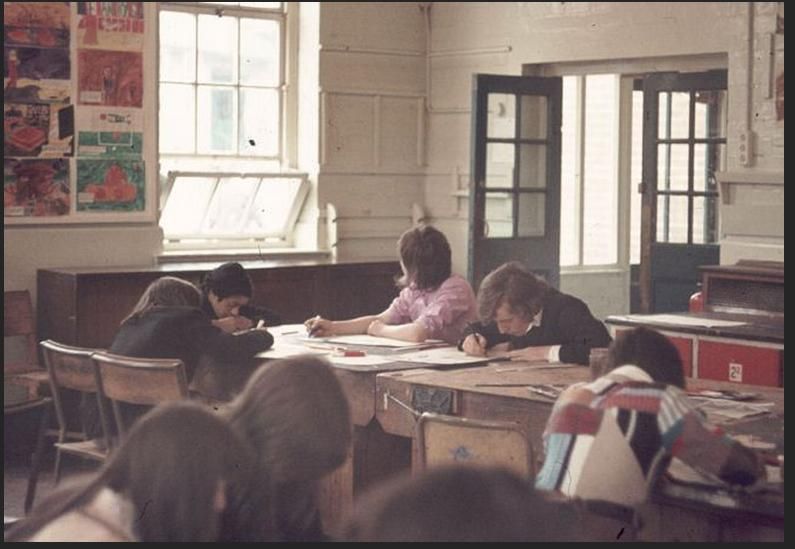 Colour photograph of teenage pupils drawing at old wooden tables in a high-ceilinged classroom with tall sash windows, half-glazed doors and children’s paintings pinned on the wall.