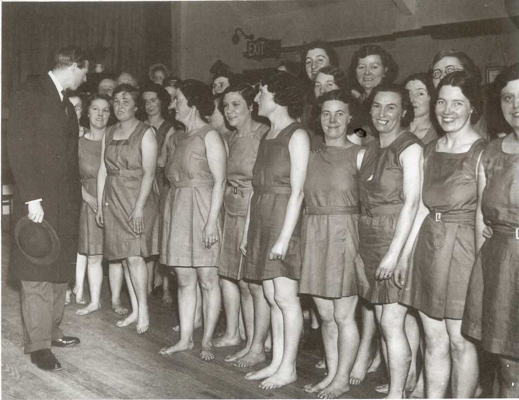 Black-and-white photograph of a line of smiling women in matching sleeveless belted tunics, barefoot on a wooden hall floor, being greeted by a man in a dark morning coat holding a hat; an EXIT sign is on the wall behind.