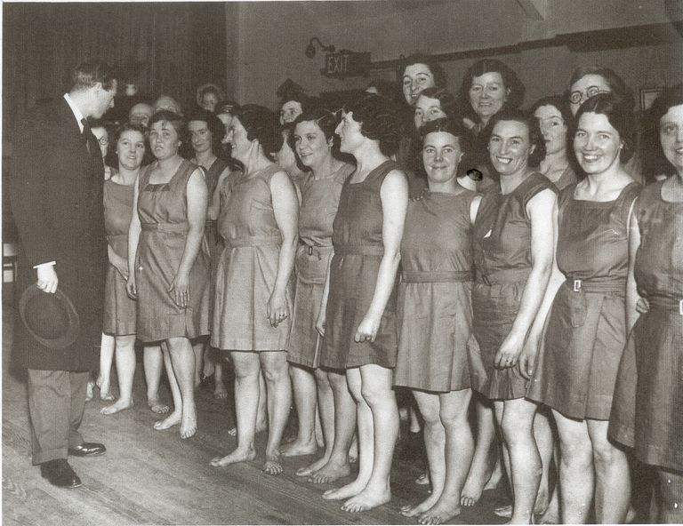 Black-and-white photograph of a line of smiling women in matching sleeveless belted tunics, barefoot on a wooden hall floor, being greeted by a man in a dark morning coat holding a hat; an EXIT sign is on the wall behind.