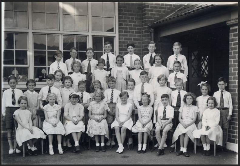 Black-and-white class photograph of about forty ten-year-old boys and girls in four rows outside a brick school building with large metal-framed windows; the boys wear ties and the girls summer frocks.