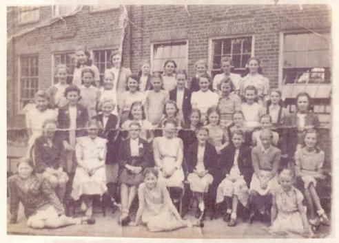 A creased class photograph of about thirty-five girls in four rows outside a brick school building, some seated on chairs and two sitting on the ground in front.