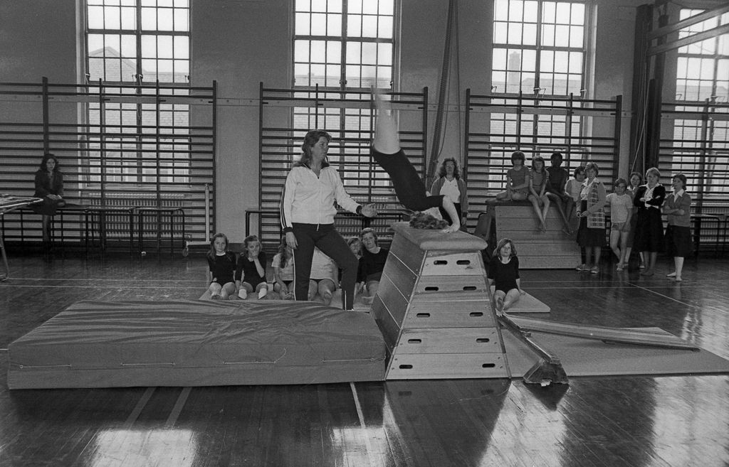 Black-and-white photo of a girl vaulting head-first over a wooden vaulting box onto a crash mat while a woman teacher in a tracksuit stands ready; girls in leotards sit on mats and others watch from wall bars beneath tall windows.