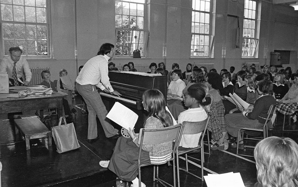 Black-and-white photo of a crowded school hall: a teacher in flared checked trousers plays an upright piano on a low stage while rows of pupils holding song sheets sing, and another teacher sorts papers at a table on the left.